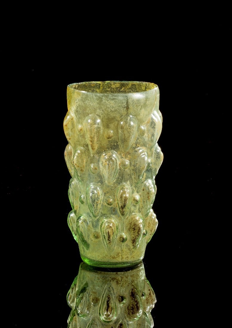 Mandelbecher.: Almond cup. Eastern Mediterranean, 1st half 1st c. AD. H 12,7cm. Greenish clear glass. Conical, mould-blown body with almond-shaped knobs. Cracks in the body, reassembled, one large piece at the rim r
