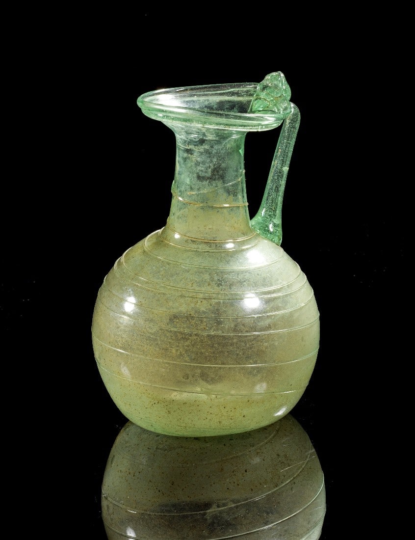 Kanne.: Pitcher with bulbous body with trail decoration, folded rim and slim handle forming a thumbrest on rim. H 12,2cm. Roman, 1st - 3rd century AD. Green translucent glass. Light iridescence, two cracks on
