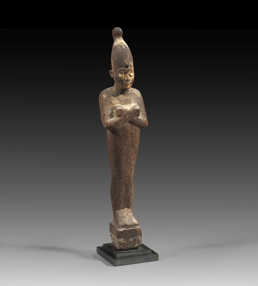 Osiris.: Osiris. Dritte Zwischenzeit, 25. Dynastie, ca. 700 v. Chr. H 27,8cm. Holz. Mumienförmige Statuette mit überkreuzten Armen auf Basis. Die Augen in Knochen eingelegt, um die Augen blaue Farbreste.