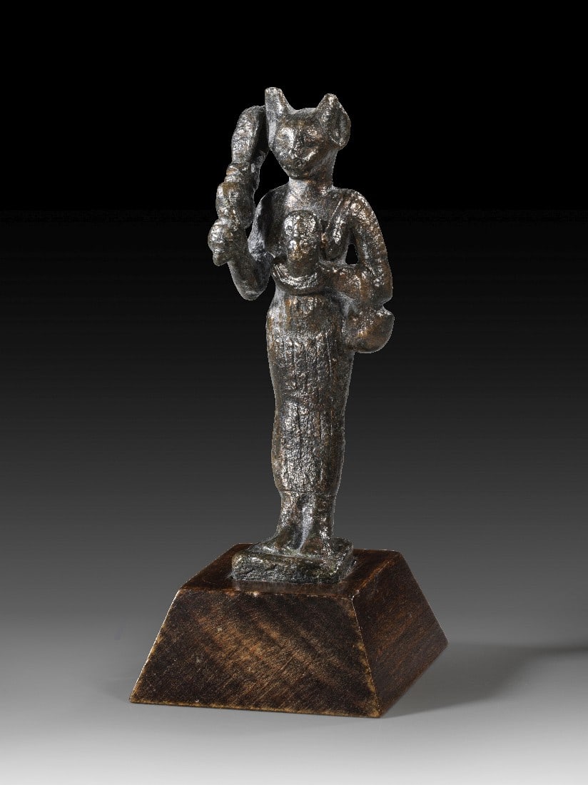 Bronzestatuette der Bastet.: Bronzestatuette der Bastet. 3. Zwischenzeit bis Spätzeit, ca. 1070 - 664 v. Chr. H 8,7cm, mit Holzsockel 10,5cm. Bronzevollguss. Stehende weibliche Statuette mit eng anliegendem Gewand und