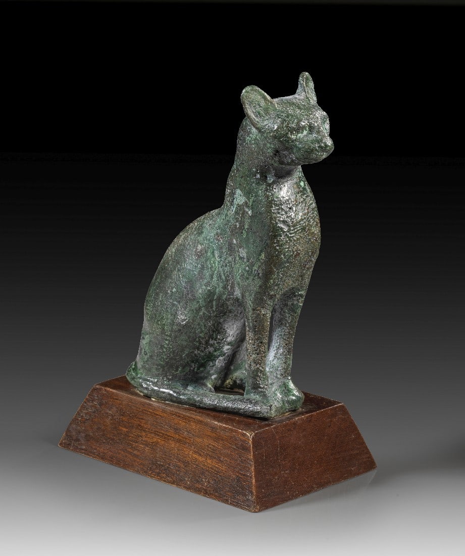 Bronzestatuette der Bastet.: Bronzestatuette der Bastet. 3. Zwischenzeit bis Spätzeit, ca. 1070 - 664 v. Chr. H 13cm, mit Holzsockel 16cm. Bronzehohlguss. Aufrecht sitzende Katze, der Schwanz um den Körper gelegt. Dargestellt