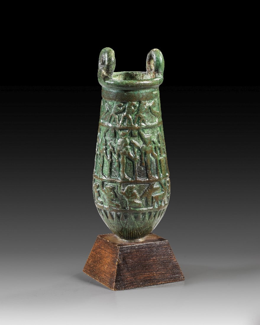 Bronze-Situla mit Reliefdekor.: Bronze-Situla mit Reliefdekor. Ptolemäisch, ca. 305 - 30 v. Chr. H 12cm, mit Holzsockel 14,5cm. Bronzevollguss. Am oberen Rand zwei Ösen für die Aufnahme des Henkels. Corpus mit drei Registern mit