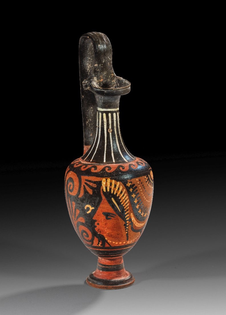 Schlanke apulische rotfigurige Oinochoe der Kantharos-Gruppe.: Schlanke apulische rotfigurige Oinochoe der Kantharos-Gruppe. 320 - 310 v. Chr. H 28,5cm, ø 9,9cm. Kanne des Typus 1 mit Kleeblattmündung und hohem Bügelhenkel. Auf der Vorderseite Frauenkopf mit