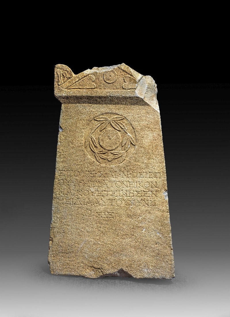 Grabstein mit Giebel, Lorbeerkranz und griechischer Inschrift.: Grabstein mit Giebel, Lorbeerkranz und griechischer Inschrift. Römisch, östliches Mittelmeergebiet, 2. - 3. Jh. n. Chr. H 57cm, B unten 35cm, T 8cm. Marmor. Platte, deren Längskanten leicht