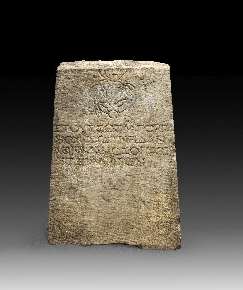 Grabstele mit Lorbeerkranz und griechischer Inschrift.: Grabstele mit Lorbeerkranz und griechischer Inschrift. Römisch, östliches Mittelmeergebiet, 2. - 3. Jh. n. Chr. H 37,5cm, B unten 35cm, T 7,5cm. Weißer Marmor. Platte, mit einem gebundenen