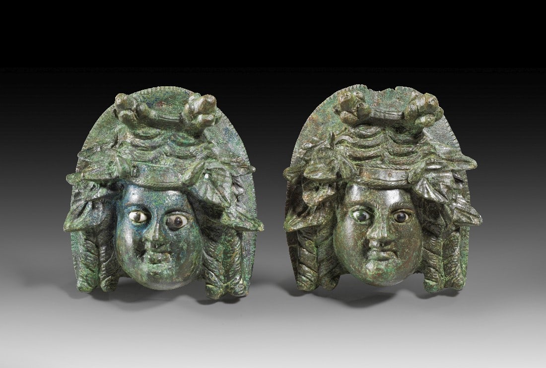 Paar Theatermasken.: Paar Theatermasken. Frühe römische Kaiserzeit, 1. Jh. n. Chr. H 7,2cm. Einseitig ausgeformte Appliken in Gestalt von Masken mit Korkenzieherlocken und großen Efeu-Korymbenkränzen. Die Augen sind