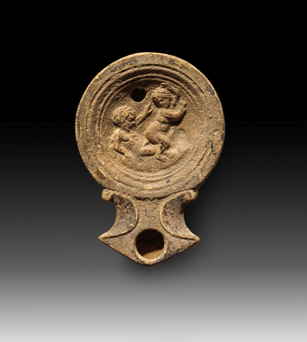 Römische Tonlampe mit Paar beim Liebesspiel.: Römische Tonlampe mit Paar beim Liebesspiel. Kaiserzeitlich, 1. - 2. Jh. n. Chr. L 9cm. Typus Loeschcke IV. Hellbeiger Ton mit braungrauem Überzug. Auf dem Spiegel eine erotische Szene à tergo.