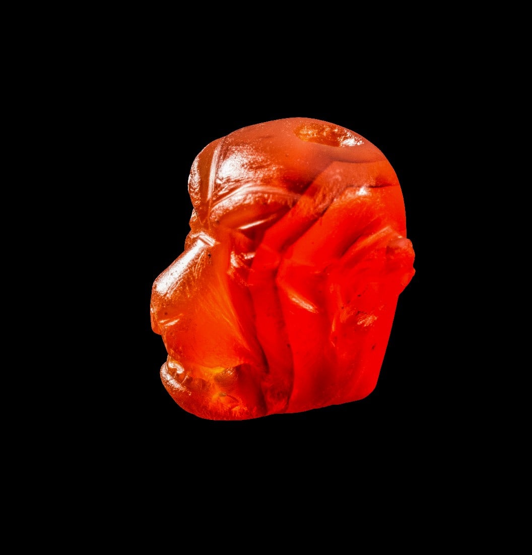 Figürlicher Karneolanhänger in Gestalt eines Pazuzu-Kopfes.: Figürlicher Karneolanhänger in Gestalt eines Pazuzu-Kopfes. Neoassyrisch, 8. - 7. Jh. v. Chr. H 1,7cm. Oranger Karneol. Hochkant durchbohrt. Fratzenhaftes Gesicht mit Hundeschnauze und spitzen