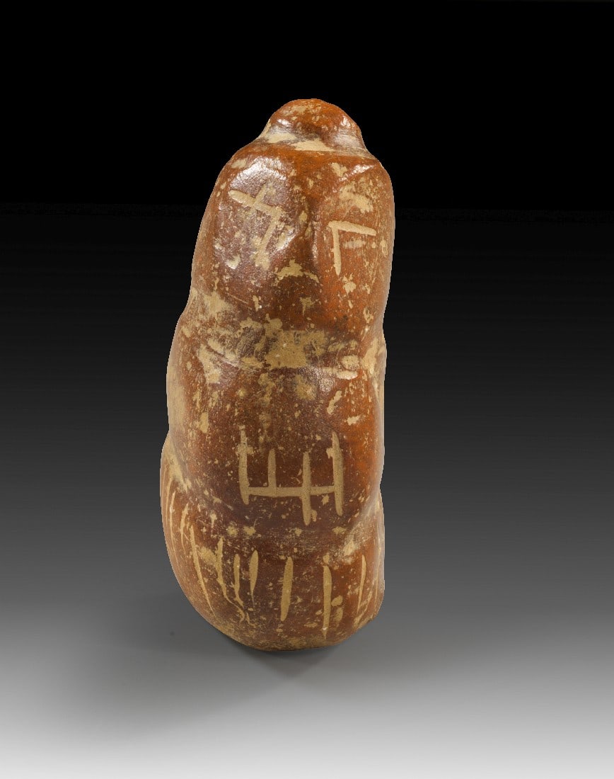 Primitives Vinca-Idol.: Primitives Vinca-Idol. Südosteuropa, 5. Jt. v. Chr. H 7,5cm. Rotbrauner Ton. Bohnenförmige Figur mit stilisiertem Gesicht. Auf dem Kopf sitzt ein Dutt, unter dem Haare hervorsprießen. Auf dem