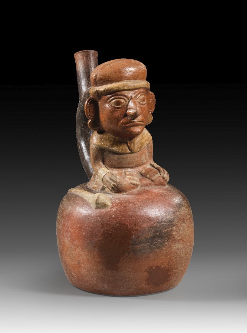 Hockender Würdenträger.: Hockender Würdenträger. Moche, ca. 300 - 500 n. Chr. H 22cm, L17cm. Grauer Ton, rötlicher Überzug und beige Bemalung. Steigbügelgefäß, auf dem ein Mann mit Haube, Scheibenohrringen und