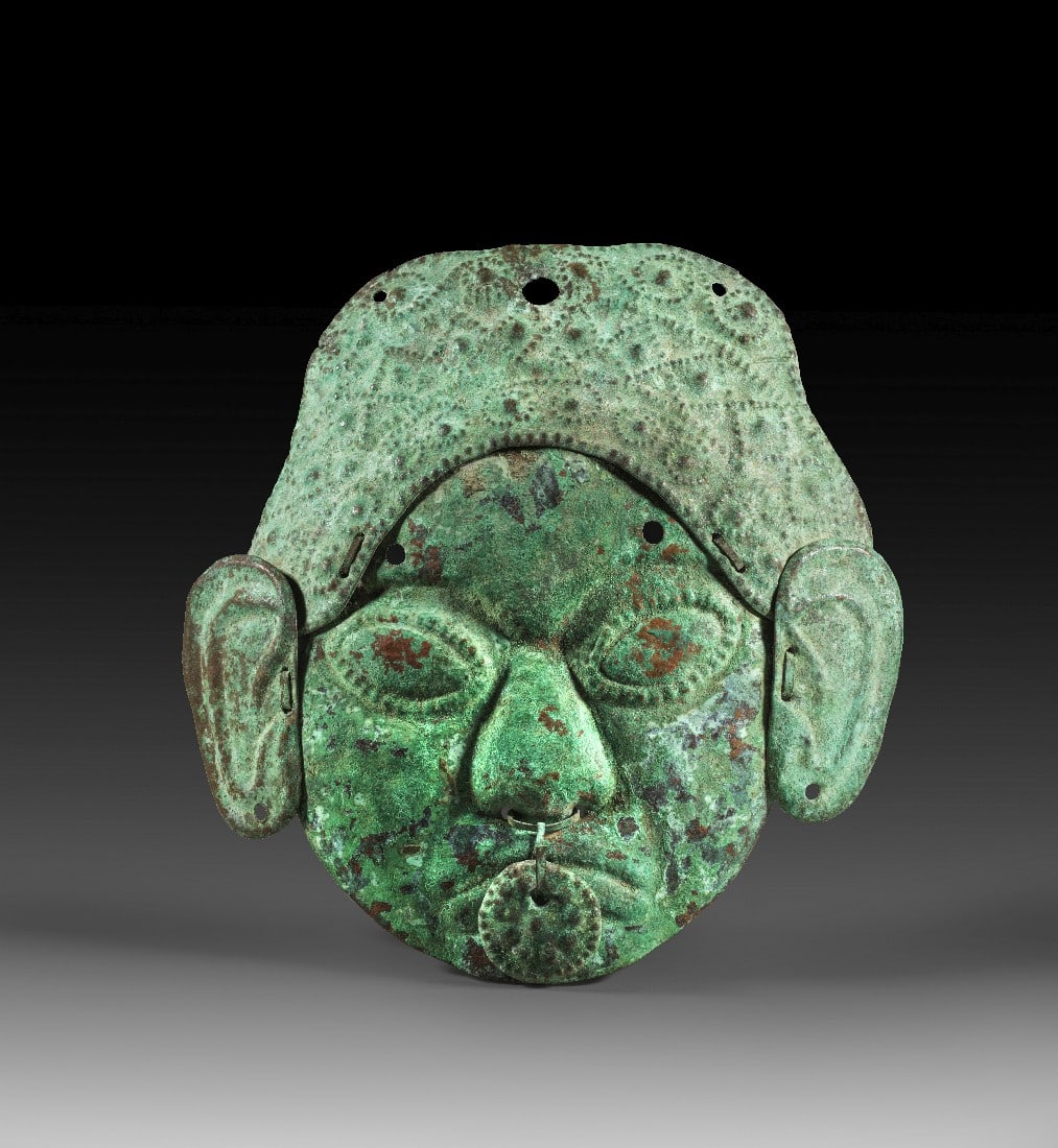 Kupfermaske.: Kupfermaske. Moche, 500 - 800 n. Chr. H 19,5cm, L 17,5cm. Totenmaske aus gehämmertem Kupfer, die den Kopf eines Adligen darstellt. Die großen Ohren und die mit geometrischen Mustern verzierte Haube