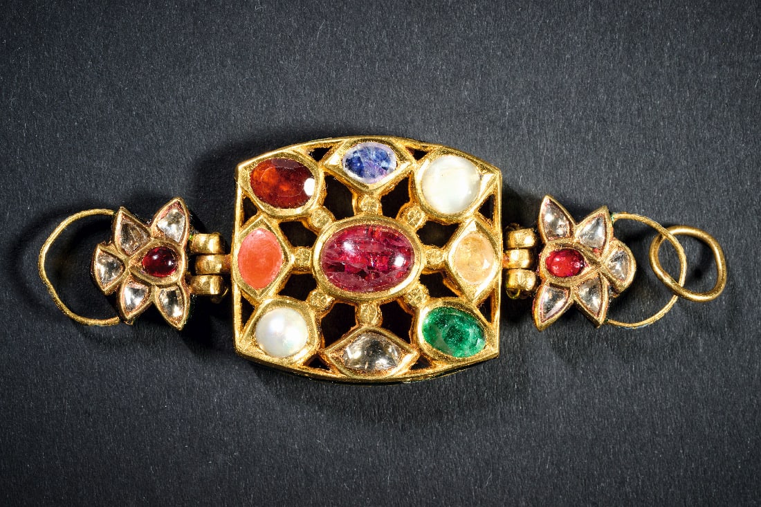 Teil eines Oberarmbandes aus Gold und Juwelen.: Teil eines Oberarmbandes aus Gold und Juwelen. Indien, 19. Jh. 24,66g, L 7,2cm. Rechteckiges, durchbrochen gearbeitetes Element, das auf der Vorderseite mit neun Juwelen "nou-ratna" besetzt ist und