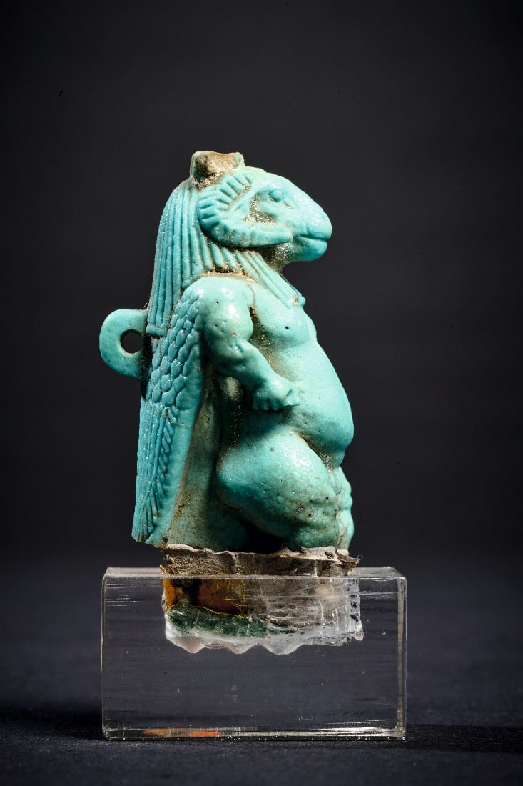 Chnum.: Chnum. Spaetzeit, 664 - 332 v. Chr. H 4,8cm. Amulett aus hellgruener Fayence. Pataeke mit Widderkopf und den Ruecken hinabfallenden Fluegeln. Krone und Fluegelspitzen gebrochen. Basis in Plexisockel
