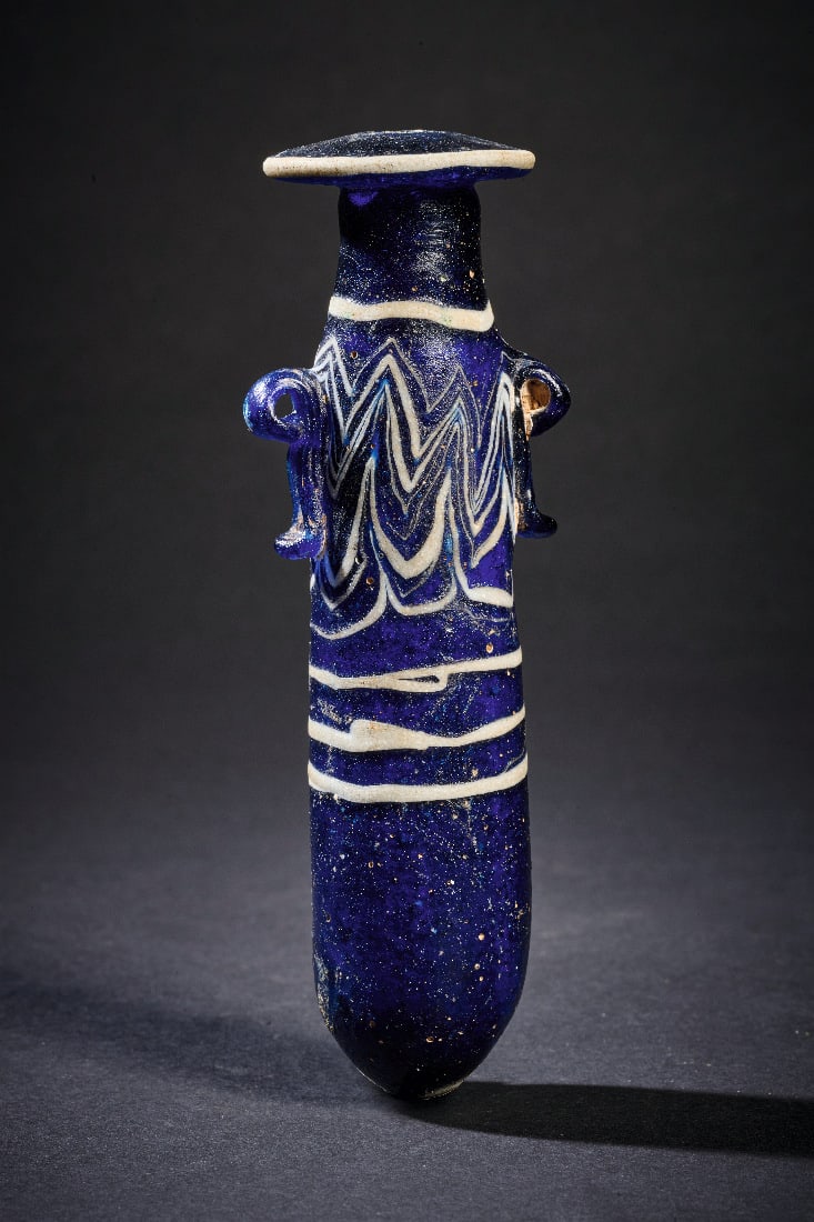 Feines Balsamarium mit weissem Zickzackdekor auf tiefblauem Grund.: Feines Balsamarium mit weissem Zickzackdekor auf tiefblauem Grund. oestliches Mittelmeergebiet, 6. - 4. Jh. v. Chr. H 12,4cm. Opak dunkelblaues Glas. Sandkerntechnik. Schlankes Balsamarium mit langges