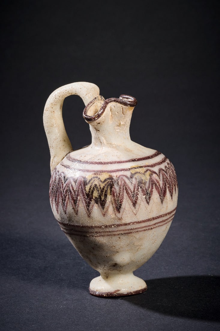 Oinochoe mit Zickzackdekor auf weissem Grund.: Oinochoe mit Zickzackdekor auf weissem Grund. oestliches Mittelmeergebiet, 6. - 5. Jh. v. Chr. H 10,2cm. Opakweisses Glas mit dunkelpurpurfarbenem Dekor. Sandkerntechnik. Kleeblattfoermig ausgezogene 