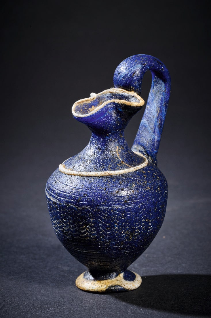 Elegante Oinochoe mit fein ausgezogenem weissem Zickzackdekor.: Elegante Oinochoe mit fein ausgezogenem weissem Zickzackdekor. oestliches Mittelmeergebiet, ca. 5. Jh. v. Chr. H 8,6cm. Glas, dunkelblau. Sandkerntechnik. Mit ausladendem Fuss, pyriformem Koerper, sch