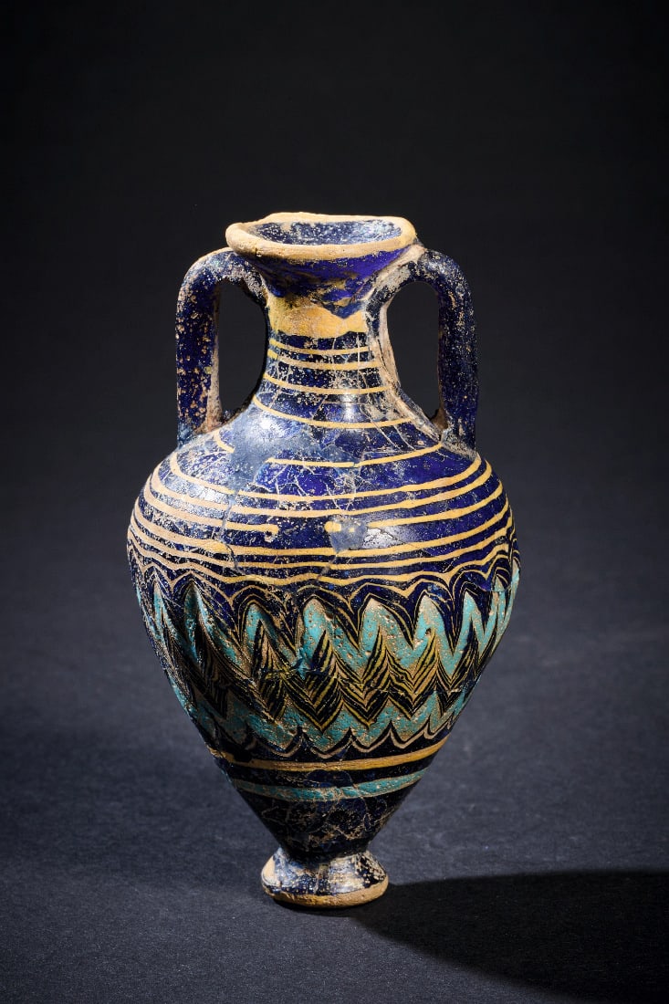 Fein dekorierter Amphoriskos mit polychromem Zickzackdekor.: Fein dekorierter Amphoriskos mit polychromem Zickzackdekor. oestliches Mittelmeergebiet, 6. - 4. Jh. v. Chr. H 9cm. Dunkelblaues, transluzides Glas. Sandkerntechnik. Eifoermiger, leicht geriefelter