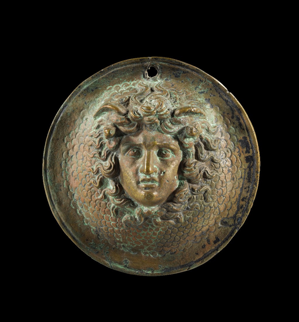 Applik mit Gorgoneion.: Applik mit Gorgoneion. Roemisch, 2. - 3. Jh. n. Chr. ? 11cm. Einseitig gestaltete Bronzescheibe mit einem frontalen Gorgoneion. Die Schuppen der aegis in Kaltarbeit. Oben Befestigungsloch.