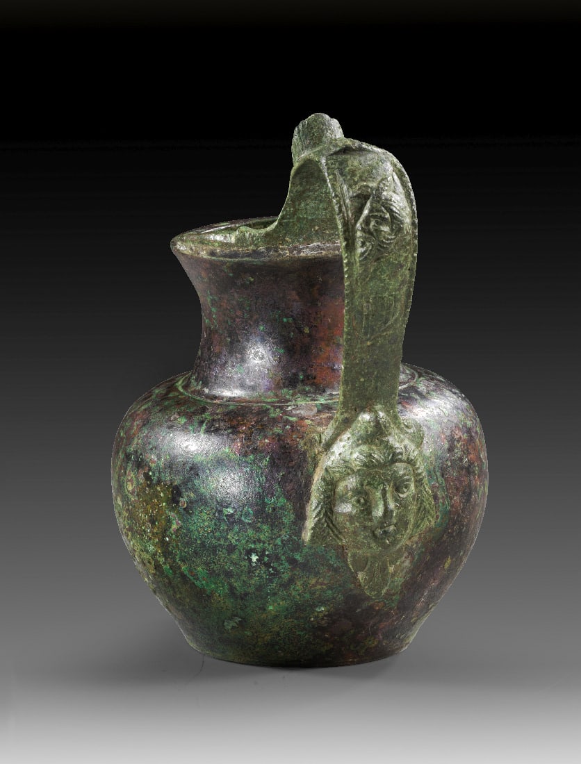 Bronzekrug mit Theatermaske.: Bronzekrug mit Theatermaske. Roemisch, 1. - 2. Jh n. Chr. H 12cm. Kleiner Krug mit bauchigem Corpus, kurzem, zylindrischem Hals und ausschwingender Muendung. Als Henkelattasche eine Theatermaske mit P