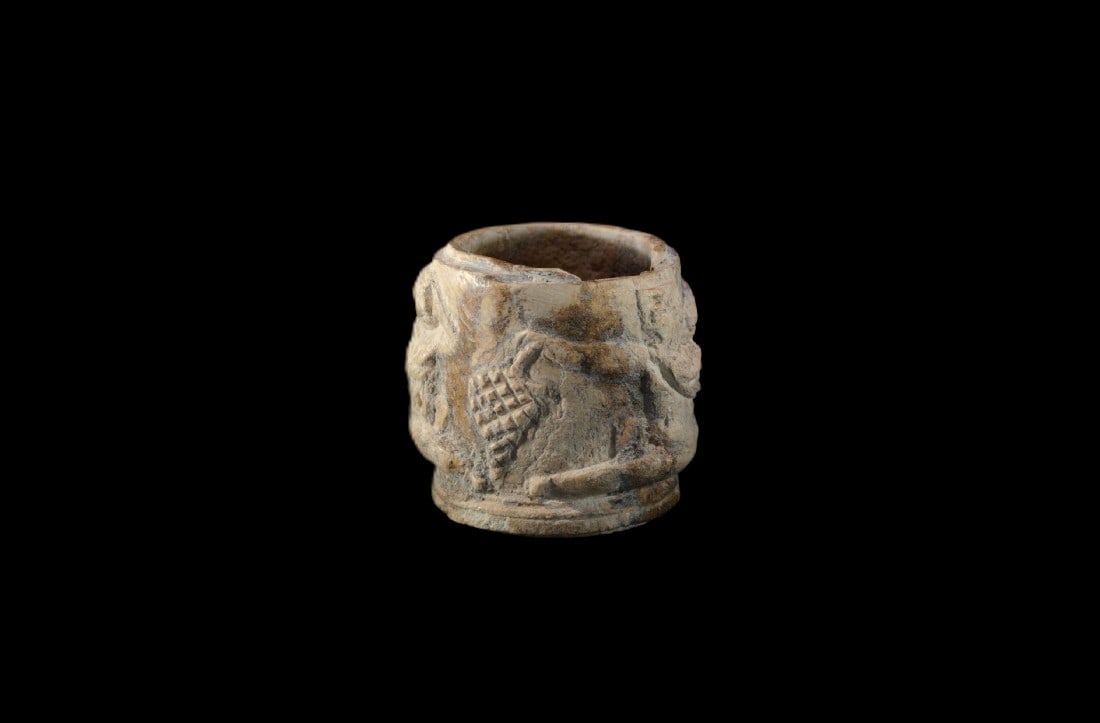 Pyxis mit Eroten.: Pyxis mit Eroten. Roemisch, 1. Jh. n. Chr. H 4cm, ? 3,7cm. Bein. Zylinderfoermig, beschnitzt mit zwei sitzenden Eroten mit Weintrauben. Auf dem Boden, der separat gearbeitet und dann eingefuegt