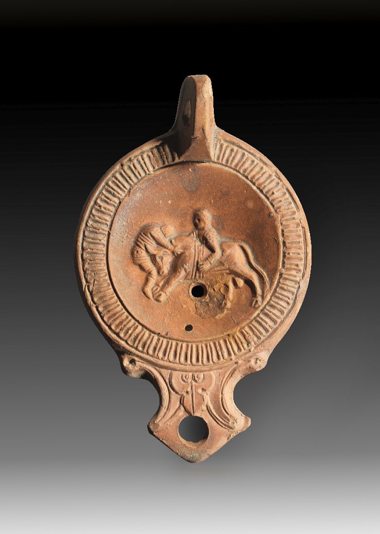 Tonlampe mit Tierkampfszene.: Tonlampe mit Tierkampfszene. Roemisch, 1. Jh. n. Chr. Loeschcke Typ IC. L 15cm, H 3,5cm. Aus roetlichem Ton mit hellrotem ueberzug. Runder Corpus mit eckiger Volutenschnauze mit rundem Brennloch und G