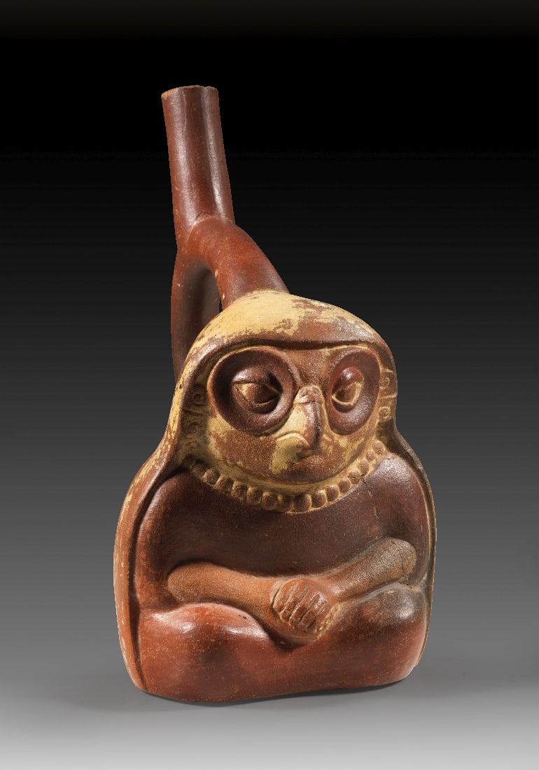 Heilerin mit Eulenmaske.: Heilerin mit Eulenmaske. Moche, ca. 800 n. Chr. H 20,5 cm, B 17 cm. Ton mit rotbraunem ueberzug. Steigbuegelgefaess in Form einer sitzenden Frau, die die Haende im Schoss gefaltet hat und eine