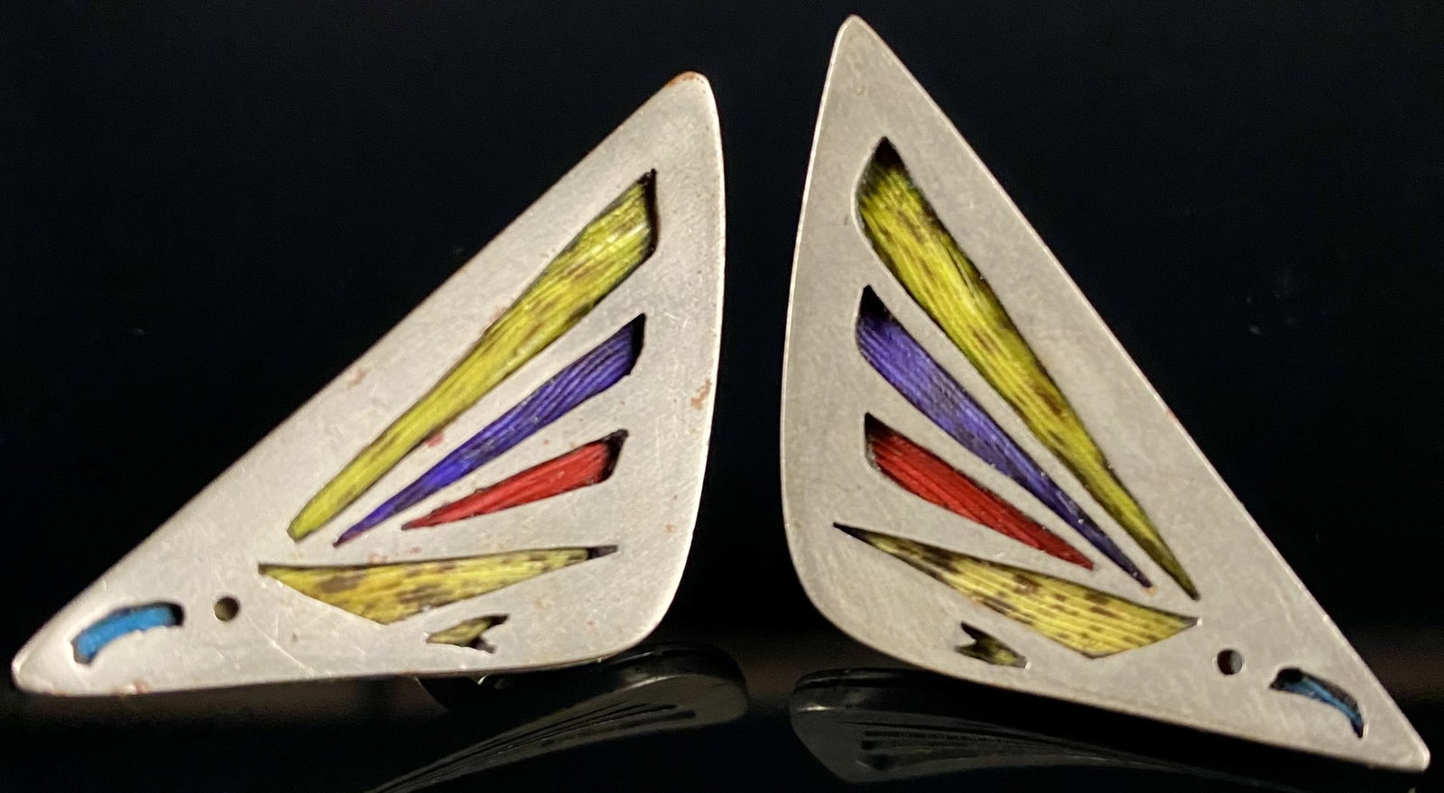 A Pair of Mexican Modernist Sterling Silver and Enamel Screw-Back Earrings: A pair of vintage screw-back earrings, each featuring a geometric triangular design crafted from sterling silver. The front of each earring is adorned with several cut-out sections filled with vibrant