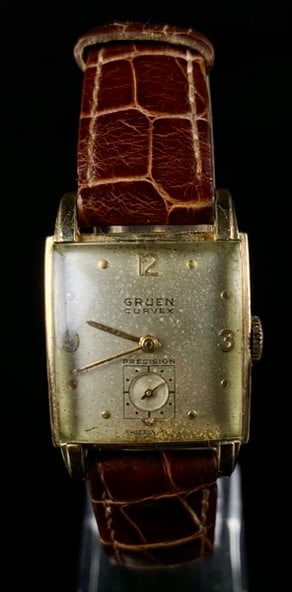 Gruen Curvex Precision 10K Gold-Filled Men's Wristwatch: A Gruen Curvex Precision 10K gold-filled men's wristwatch, featuring a rectangular, curved case. The light-colored dial presents Arabic numerals at 12, 3, and 9 o'clock, with dot markers for other hou