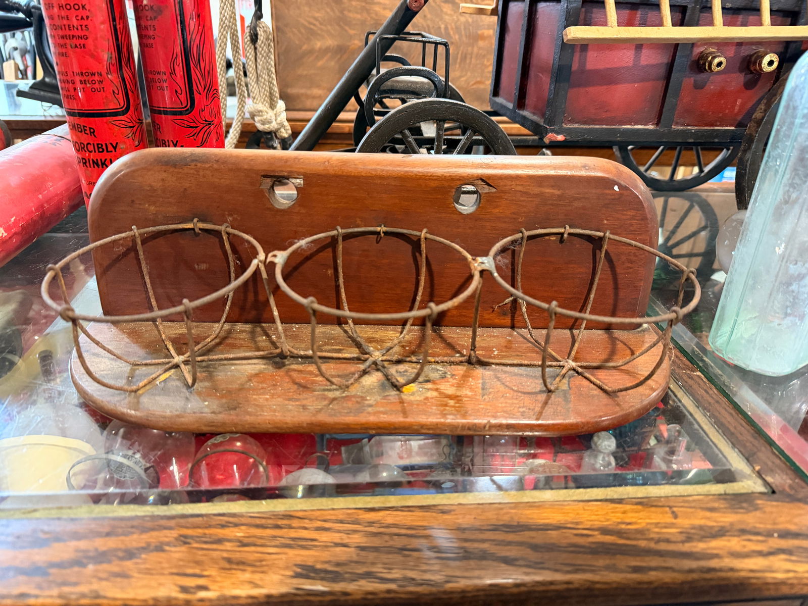 Wall Mount Fire Grenade Holder: A vintage wall-mounted or shelf-standing holder, constructed from stained wood with three semi-circular metal wire baskets for holding fire grenades. The wooden component features a rounded top edge a