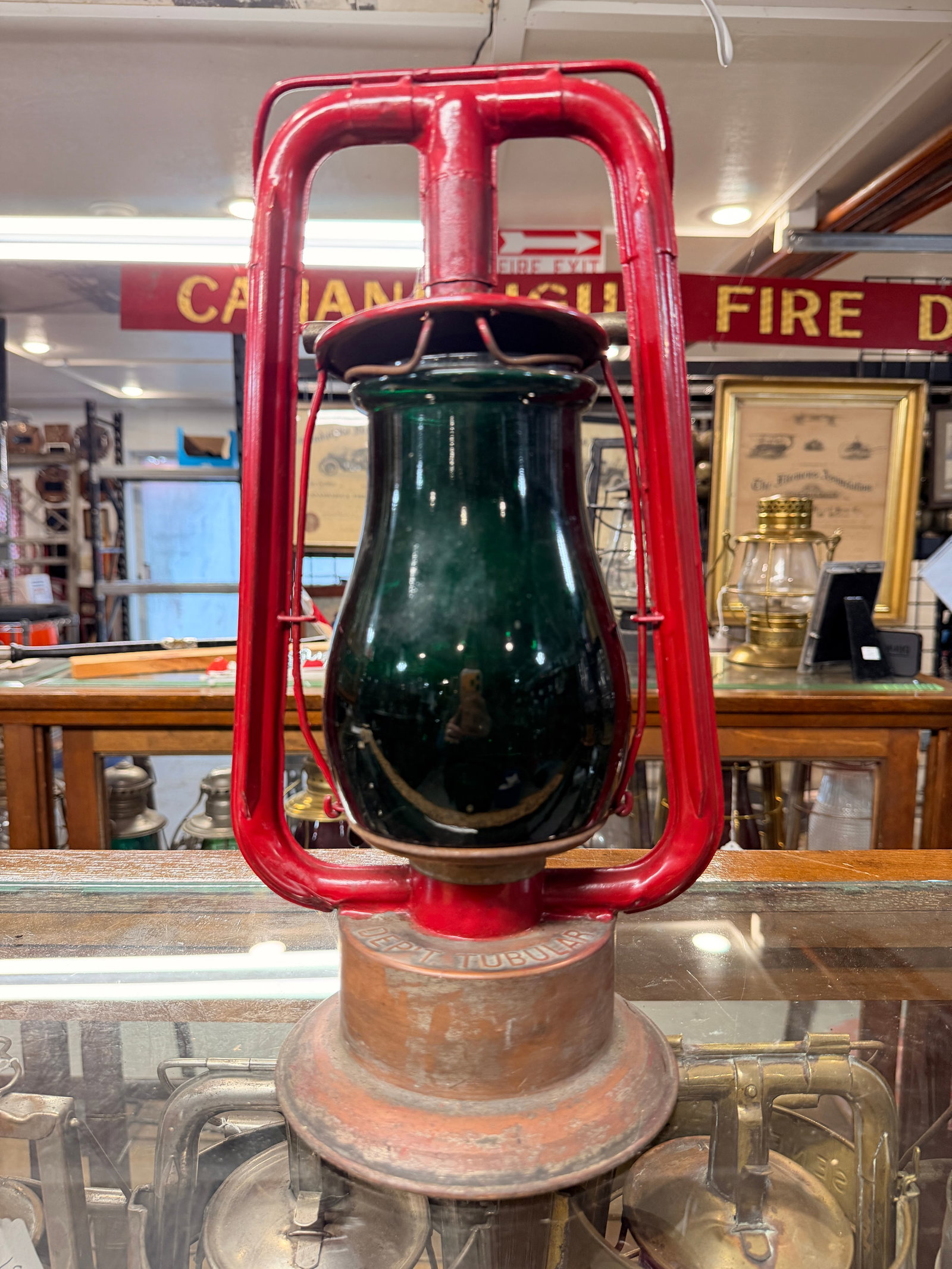Vintage Dietz Fire Tubular Signal Lantern with Green Globe: A vintage Dietz Monarch Fire tubular signal lantern, featuring a robust red-painted metal frame and a dark green glass globe. The unpainted metal base, likely copper or brass, is visibly embossed with