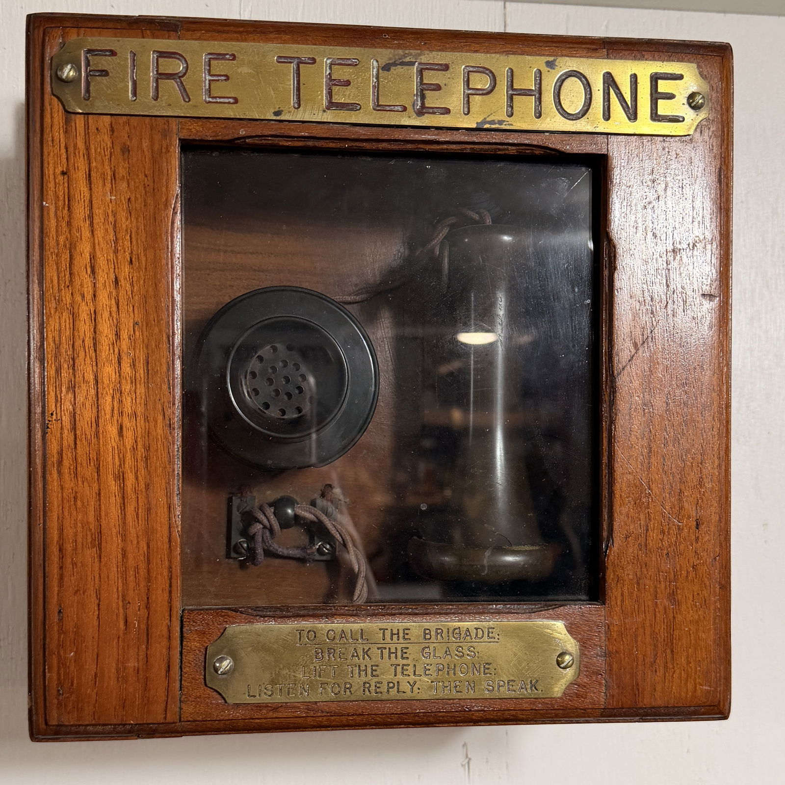 Early 20th Century Wall-Mounted 'Fire Brigade Telephone' in Oak Cabinet: A vintage wall-mounted "Fire Telephone" apparatus, housed within a rectangular polished wooden cabinet, likely constructed from oak, featuring a glass viewing panel on its front. The design and