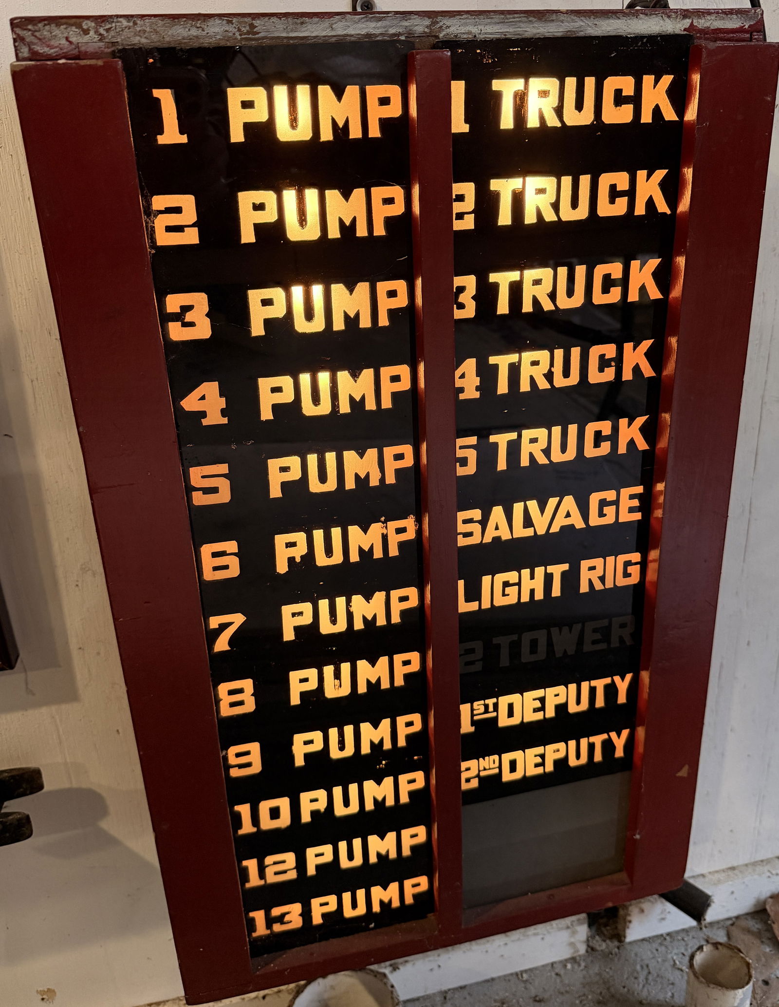 Vintage Fire Department Illuminated Status Board: A vintage illuminated display or status board, likely from a fire department, featuring a dark-red painted wooden cabinet. The translucent front panel illuminates two columns of white/yellow text on