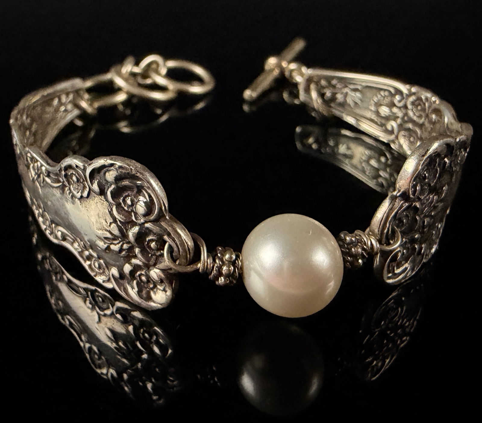 English Lace Silver and White Pearl Spoon Bracelet: This bracelet is constructed from two repurposed sterling silver flatware handles in the English Lace pattern. The metal components feature an ornate Victorian-style floral repousse design depicting r
