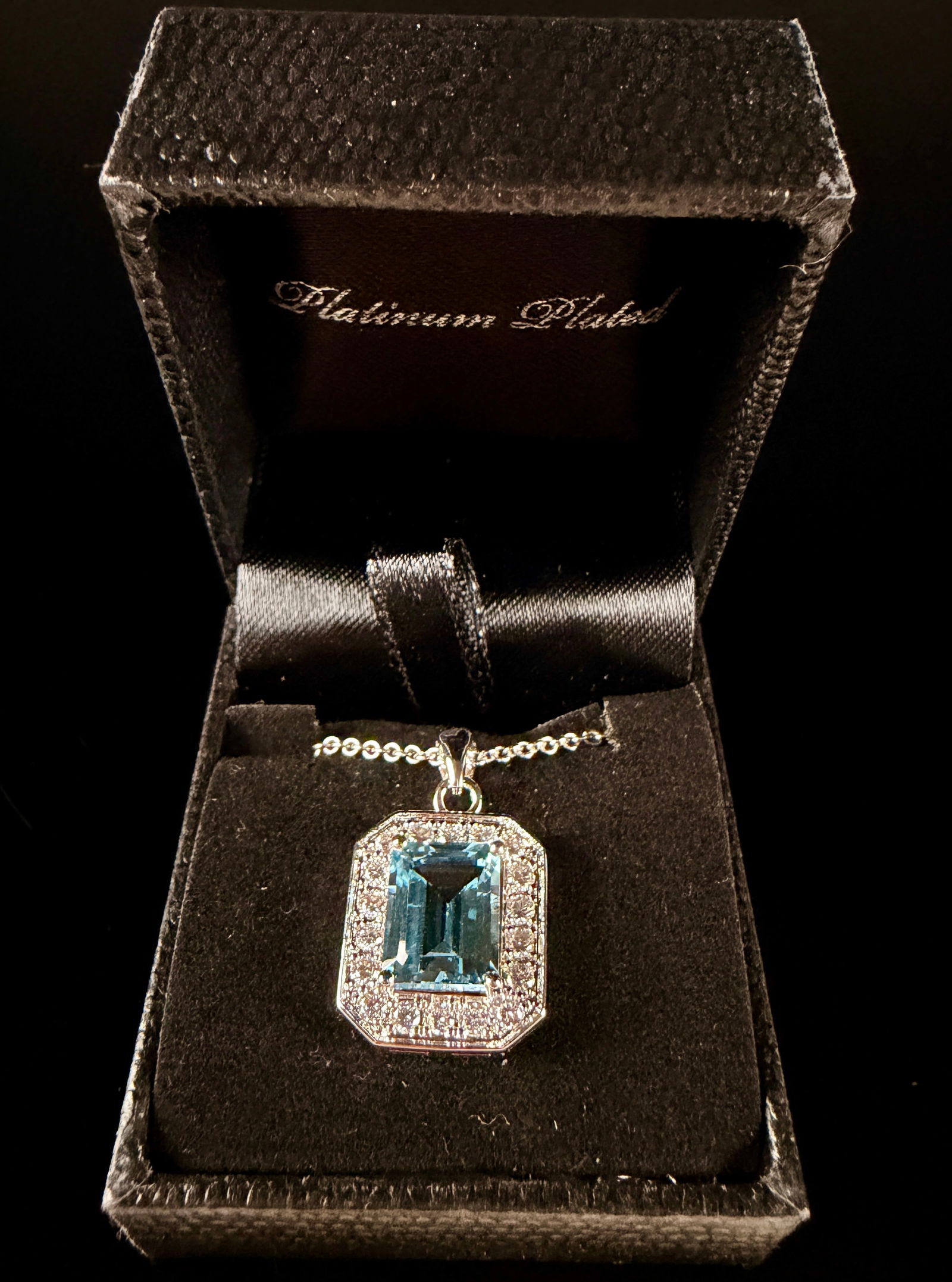 Gold Coast Platinum-Plated Blue and Clear Stone Pendant Necklace: A Gold Coast pendant necklace featuring a large rectangular emerald-cut blue stone. The central stone is bordered by a halo of round-cut clear accent stones. The pendant is suspended from a silver-ton