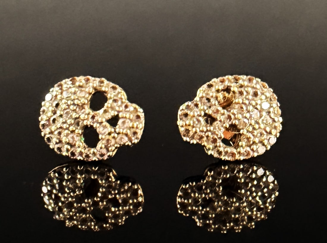 A Pair of 14k Yellow Gold and Pave-Set Stone Skull Stud Earrings: A pair of skull-motif stud earrings crafted in 14k yellow gold. The stylized skull heads feature a textured surface pave-set with numerous small, round-cut white stones. The design incorporates openwo