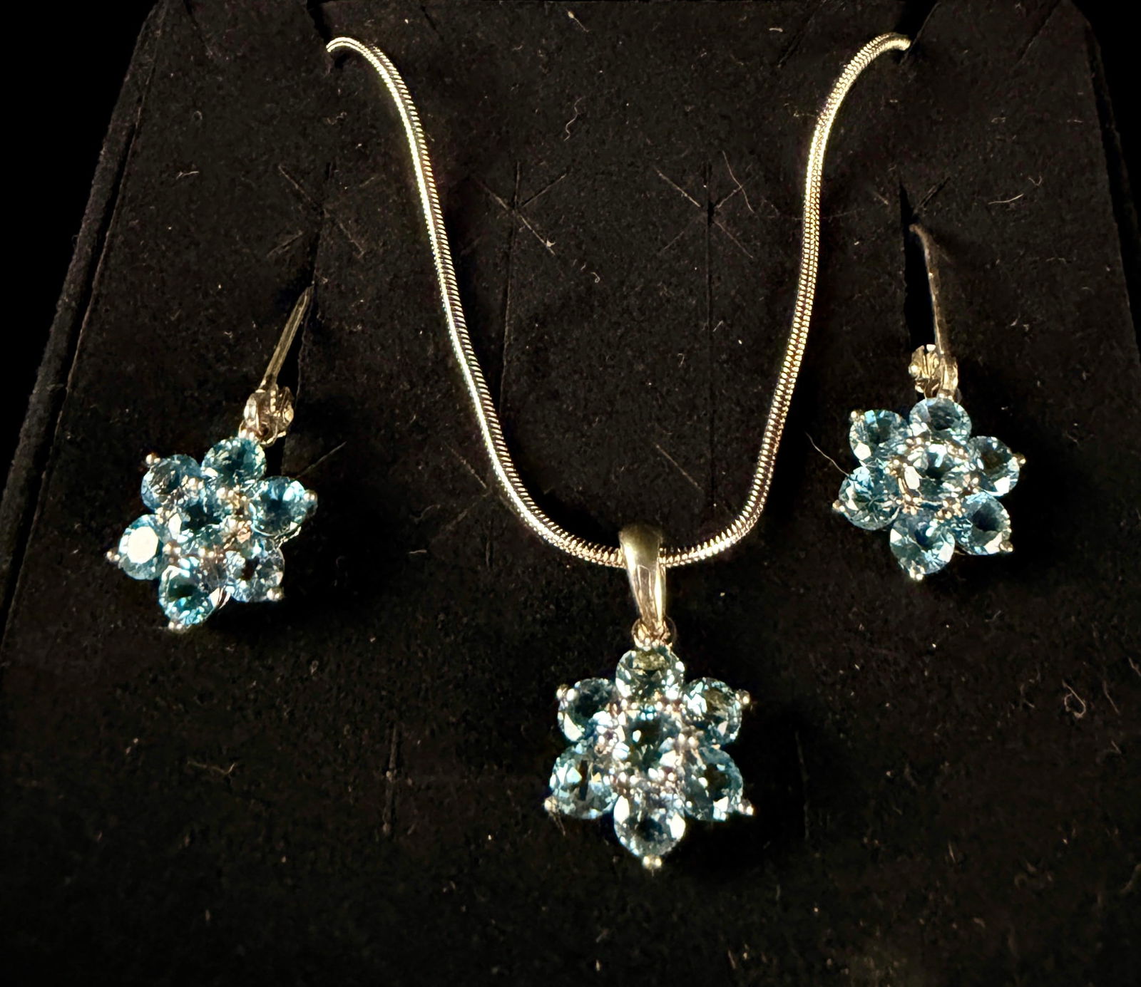 A Trio of Sterling Silver and Blue Topaz Floral Cluster Jewelry Items: A sterling silver and blue topaz jewelry set featuring a floral blossom motif. The collection includes a pendant necklace on a fine snake chain and a matching pair of drop earrings. Each piece is set