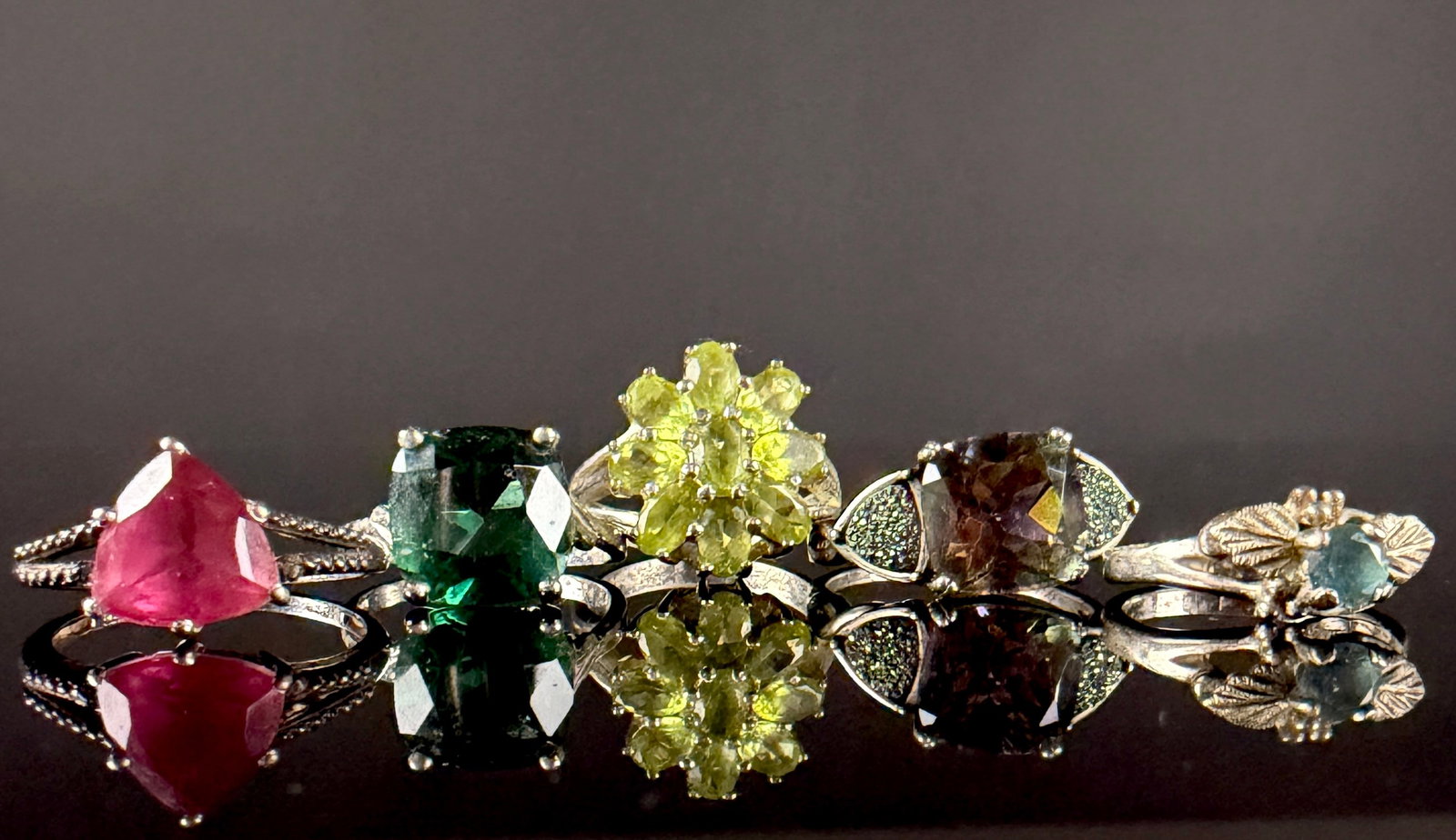 A Collection of Sterling Silver and Gemstone Rings: A collection of sterling silver rings featuring an assortment of faceted colored gemstones in various cuts and settings. The group includes a trillion-cut pink stone, a cushion-cut dark green stone, a