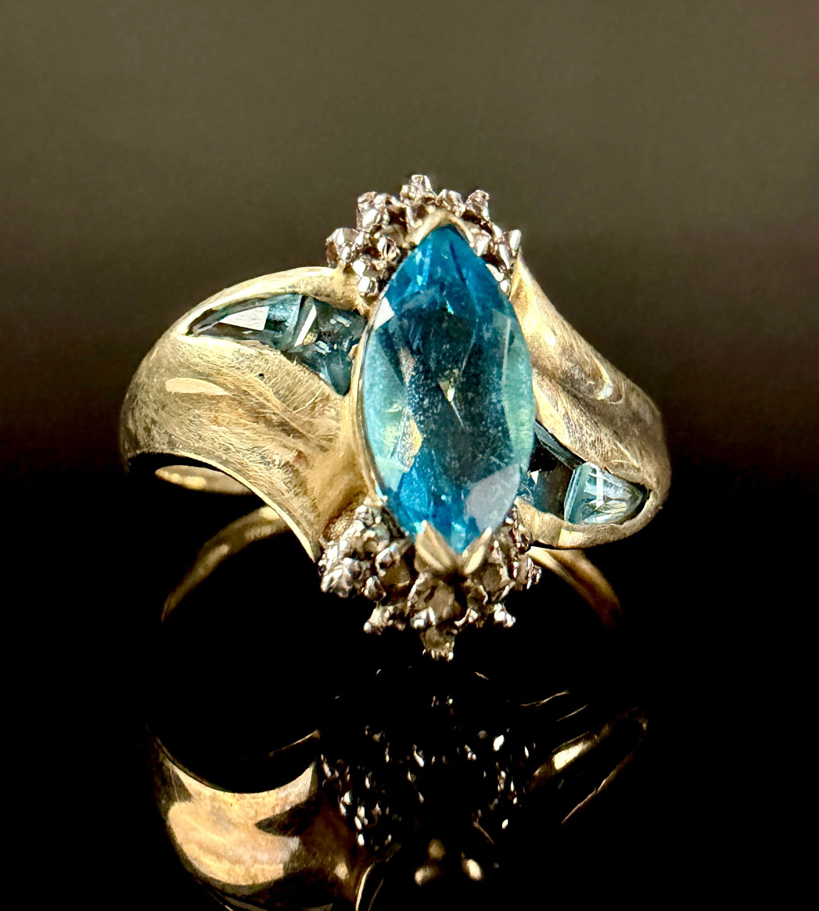 10K Yellow Gold Marquise Blue Topaz and Diamond Accent Ring: This 10K yellow gold cocktail ring features a central marquise-cut blue topaz. The primary gemstone is flanked by clusters of small diamond accents and two trillion-cut blue topaz gemstones set into t