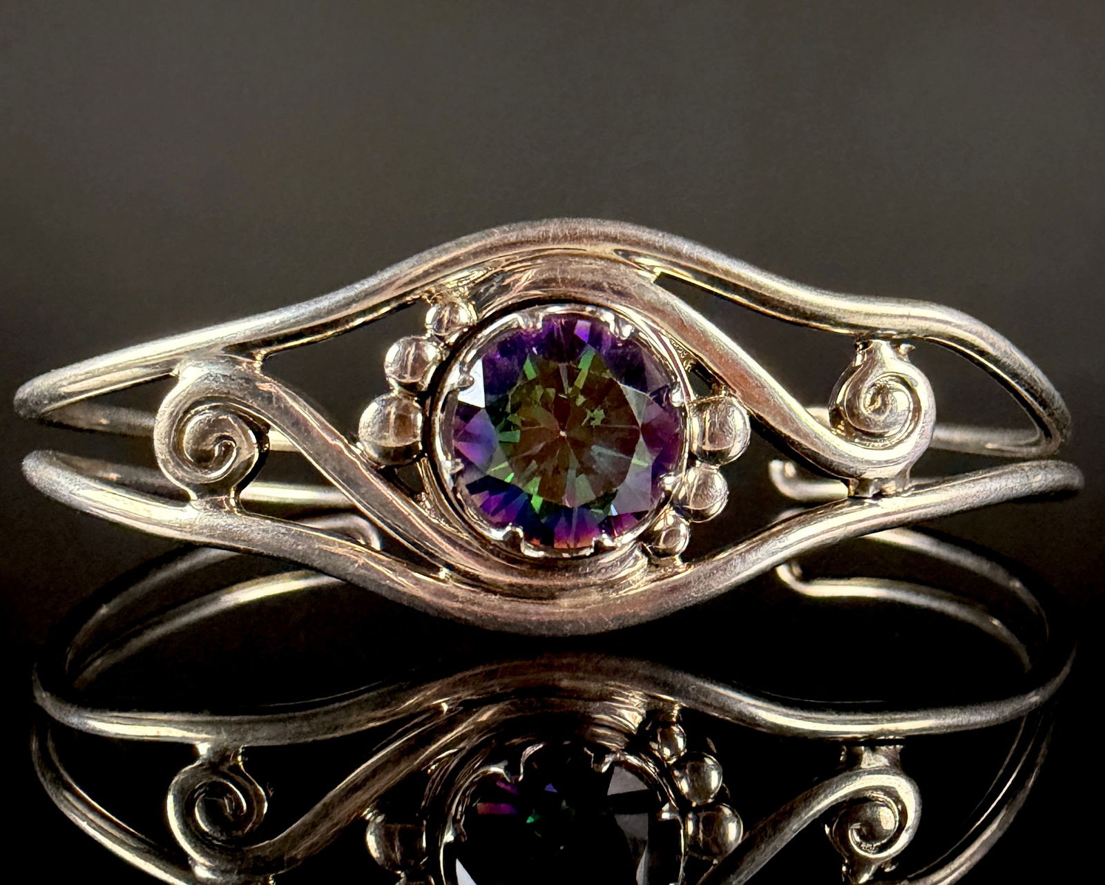 Sterling Silver and Mystic Fire Topaz Cuff Bracelet: A sterling silver cuff bracelet featuring a large round-cut faceted mystic fire topaz. The iridescent gemstone displays a spectrum of purple, green, and gold hues and is held in a multi-prong setting.