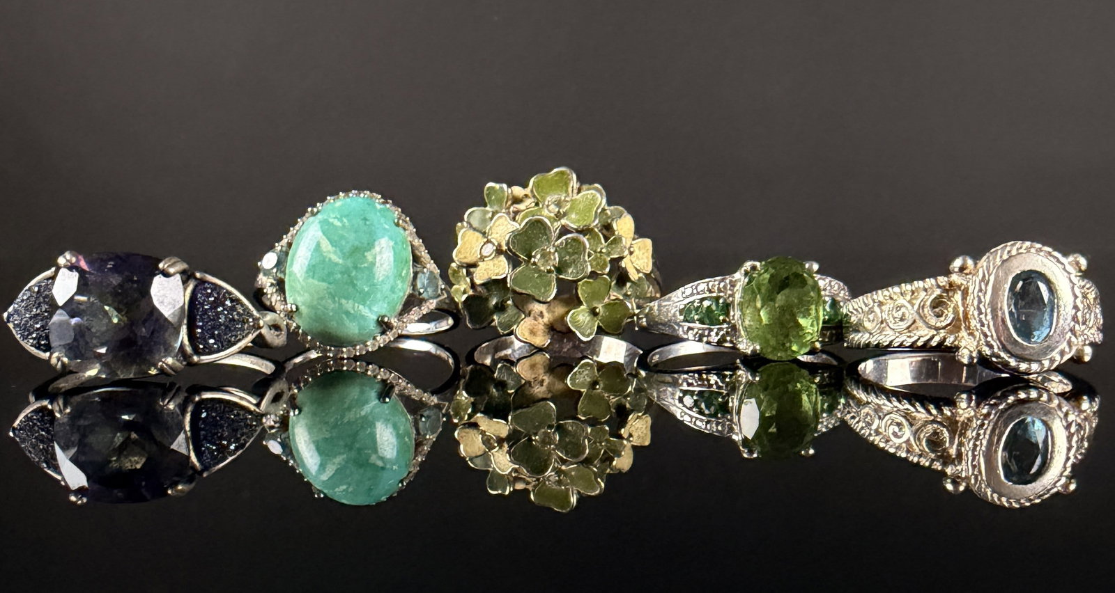 A Collection of Sterling Silver and Gemstone Rings: A collection of sterling silver rings featuring various gemstones and decorative motifs. The group includes a three-stone ring with a central faceted amethyst-tone stone and druzy-textured shoulders;