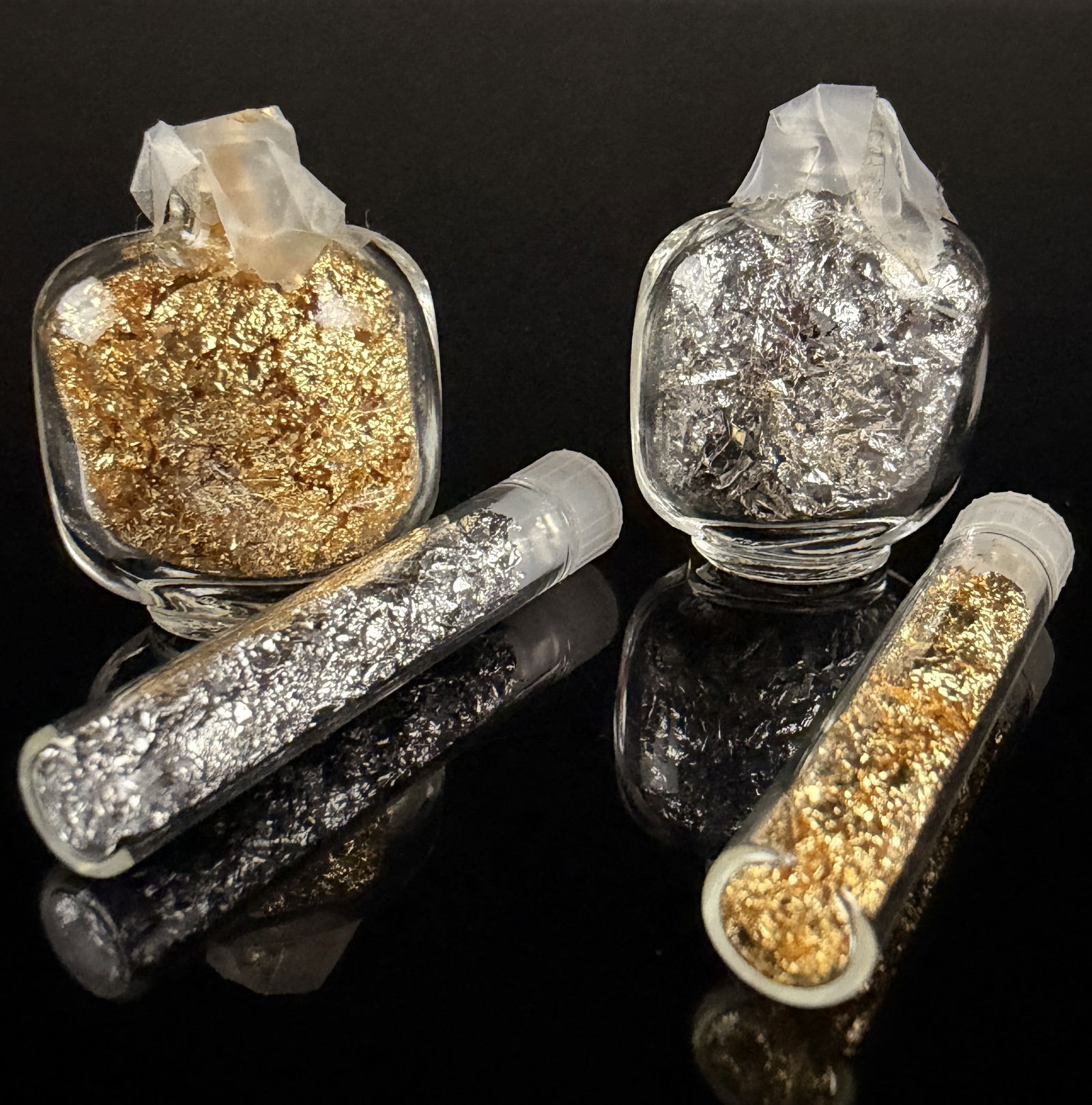 An Assortment of Gold and Silver Leaf Flakes: An assortment of gold and silvermetallic leaf flakes, commonly used for gilding, nail art, and various craft applications. The collection consists of several small, rounded glass bottles and slender g