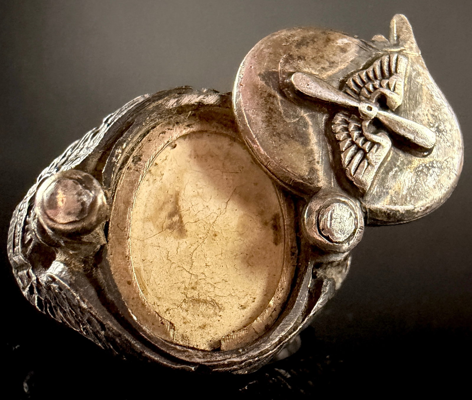 A World War II Sterling Silver USAAF Photo Locket Ring: A World War II-era sterling silver United States Army Air Forces (USAAF) sweetheart locket ring. The construction features a hinged oval face decorated with a raised winged propeller emblem that opens