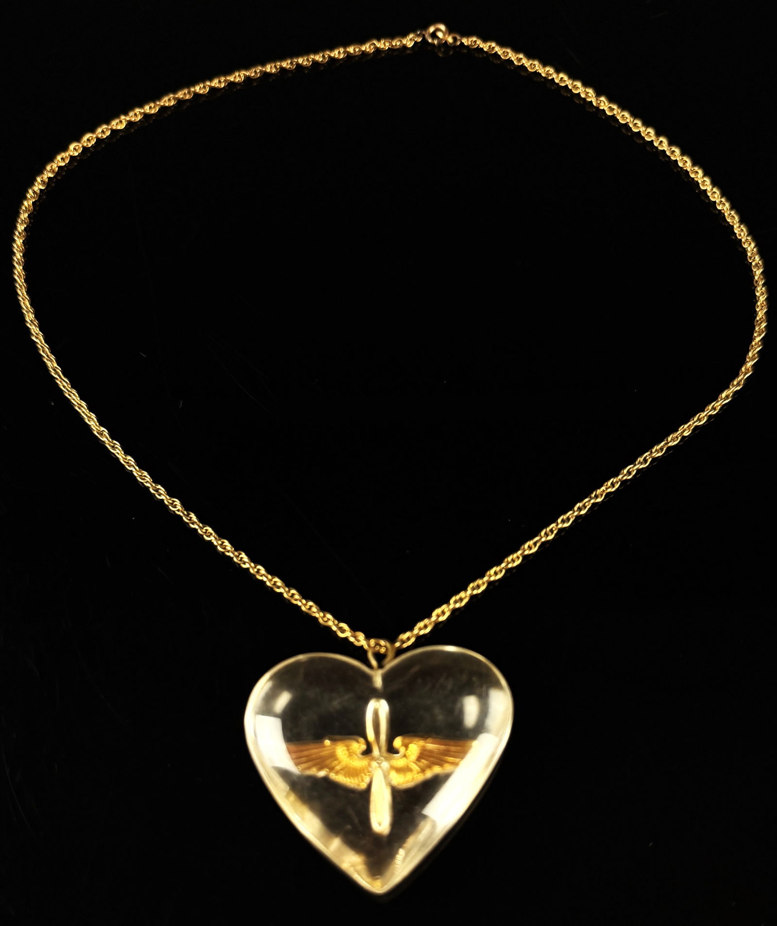 WWII US Army Air Corps Lucite Sweetheart Necklace: A WWII-era military home front sweetheart necklace featuring a clear heart-shaped Lucite pendant. The pendant contains an embedded gold-toned metal insignia of the U.S. Army Air Corps winged propeller