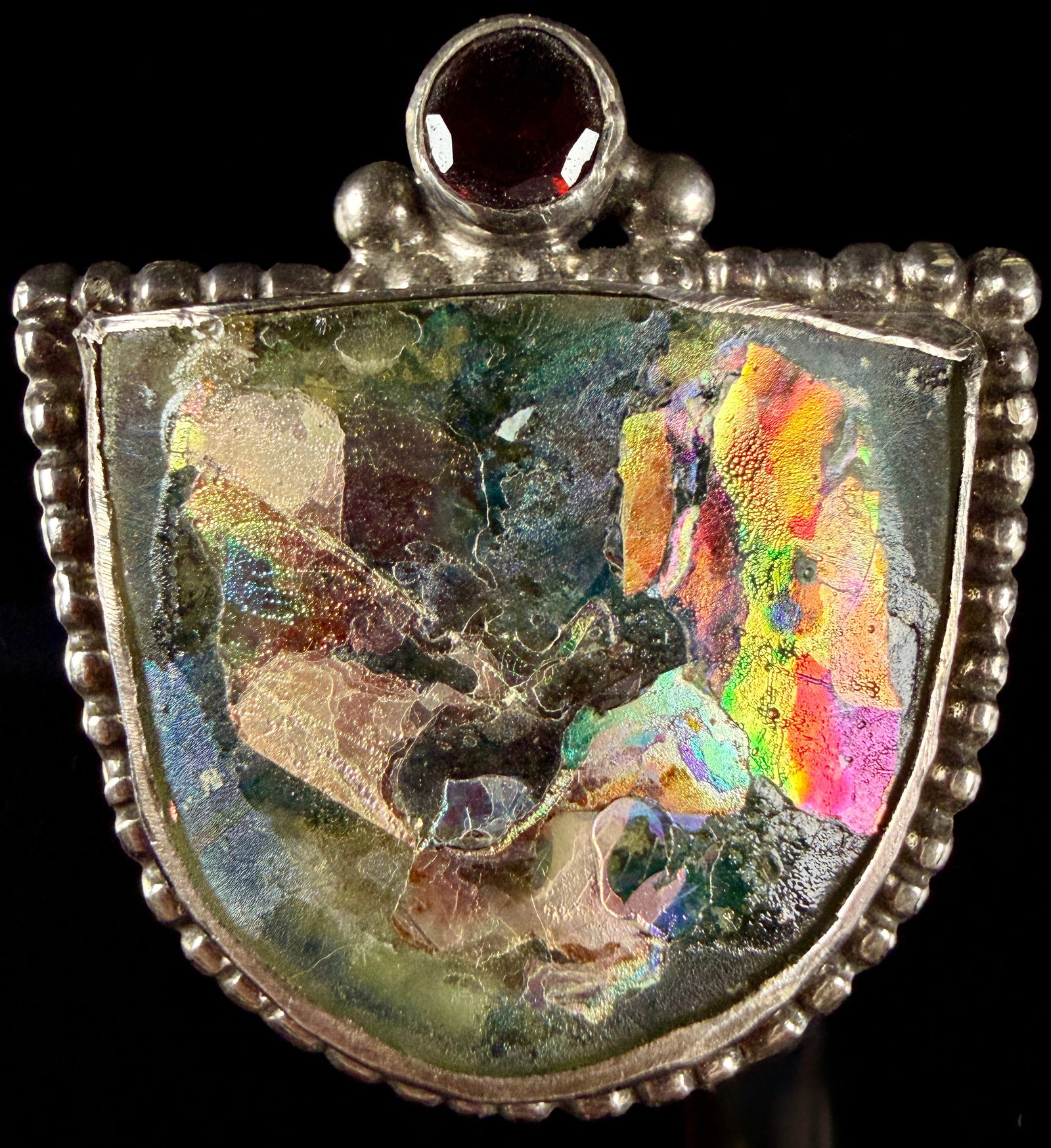 Sterling Silver Iridescent Glass and Garnet Brooch Pendant: This sterling silver jewelry piece is designed for dual use as both a brooch and a pendant. The central element is a fan-shaped fragment of iridescent glass featuring a multi-colored patina and surfac