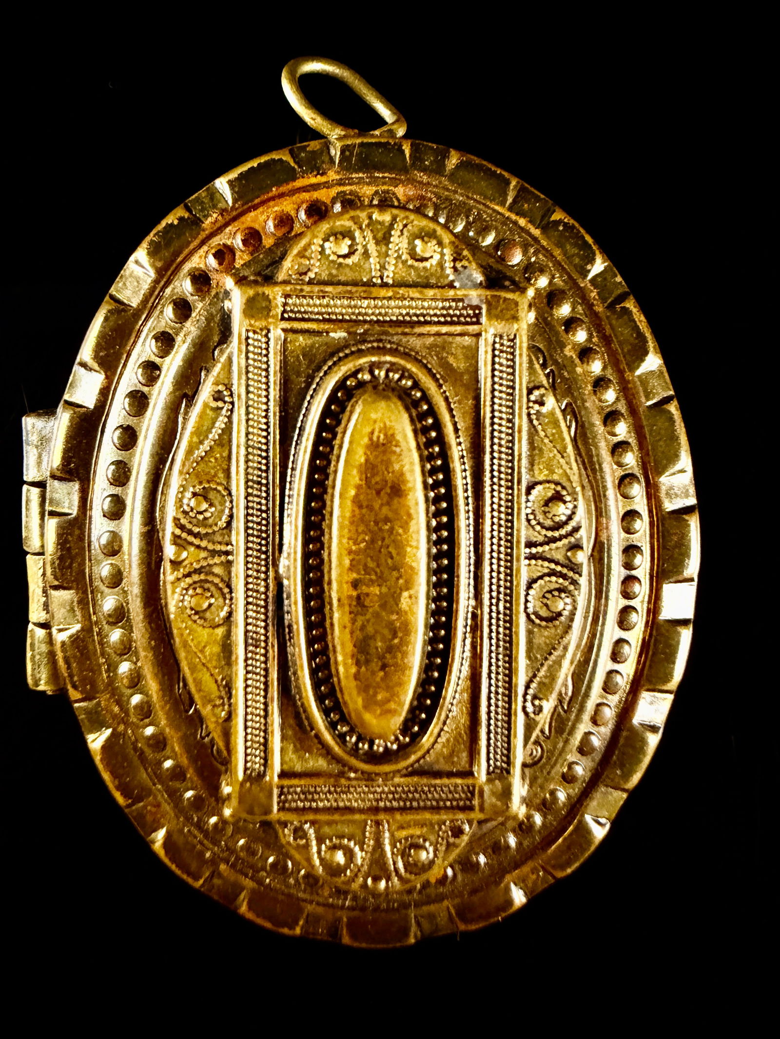 Victorian Etruscan Revival Ornate Gold Toned Oval Locket: An oval-shaped gold-toned metal locket pendant featuring Victorian Etruscan Revival styling. The exterior is decorated with ornate repousse and engraved scrollwork. The front face displays a central r