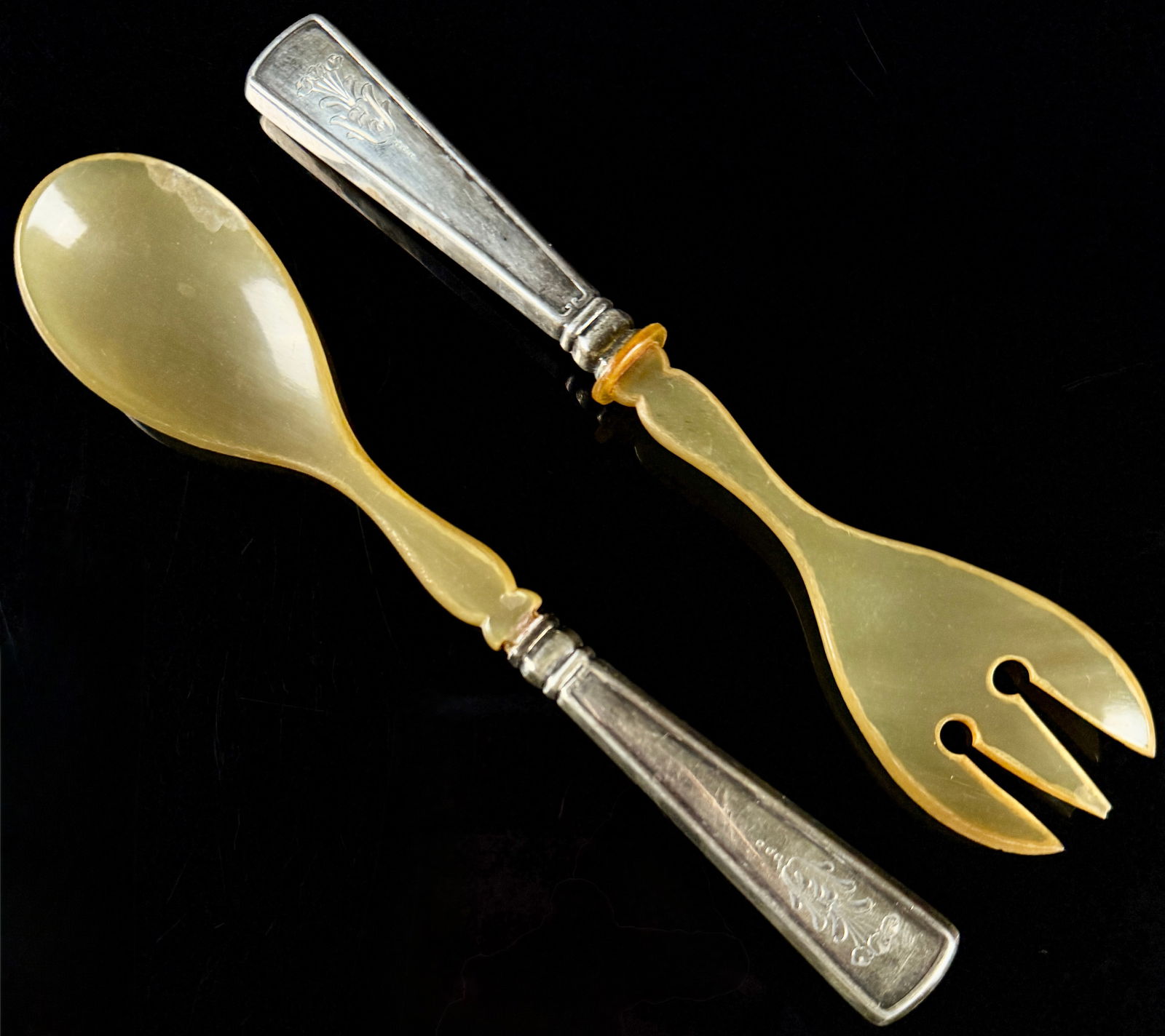 A Pair of Late 19th Century German Silver and Horn Salad Servers: A pair of antique German serving utensils consisting of a spoon and a three-pronged fork. The set is constructed with carved translucent horn heads and tapered silver handles. The handles feature pane