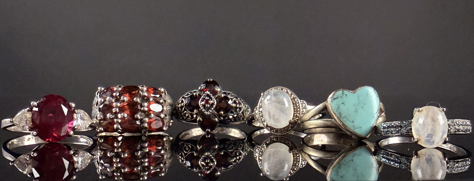A Collection of Sterling Silver and Gemstone Rings: A collection of sterling silver rings featuring a variety of gemstone settings and decorative styles. The group includes rings with faceted red stones in solitaire and cluster arrangements, a heart-sh