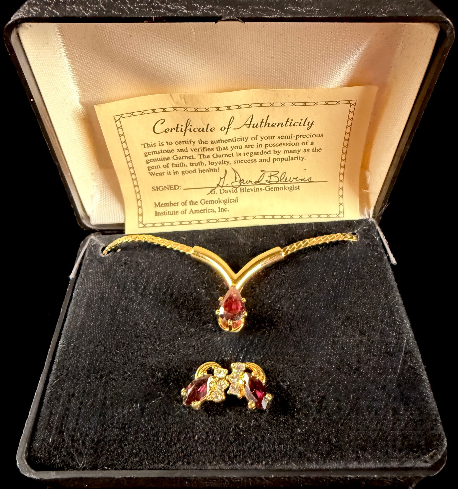 A Trio of Garnet and Gold-Toned Jewelry Including a Necklace and Matching Earrings: A jewelry suite featuring a necklace and a pair of coordinating earrings. The gold-toned necklace is constructed with a flat-link chain integrated into a chevron-shaped centerpiece. This centerpiece h