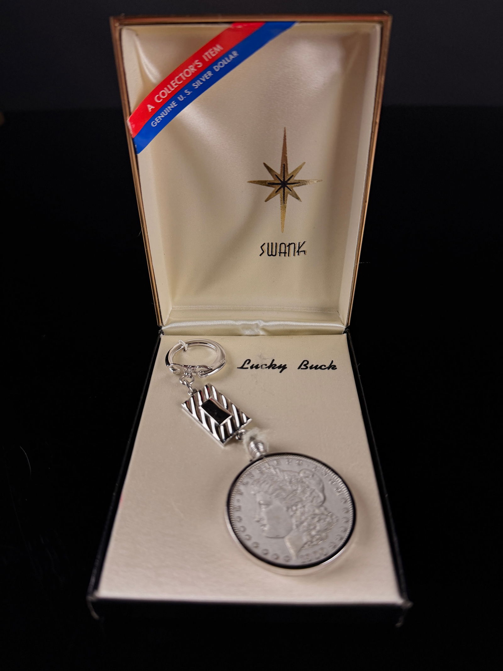 Swank Lucky Buck 1900 Morgan Silver Dollar Keychain: A vintage Swank Lucky Buck keychain featuring a genuine 1900 United States Morgan silver dollar. The coin is secured within a silver-toned bezel and connected to a decorative rectangular link