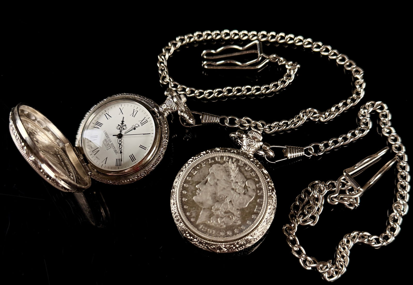 A Pair of American Historic Society Morgan Silver Dollar Quartz Pocket Watches: A pair of silver-tone hunter-case pocket watches produced by the American Historic Society, each featuring an integrated United States Morgan Silver Dollar. The coins are dated 1880 and 1891. Each